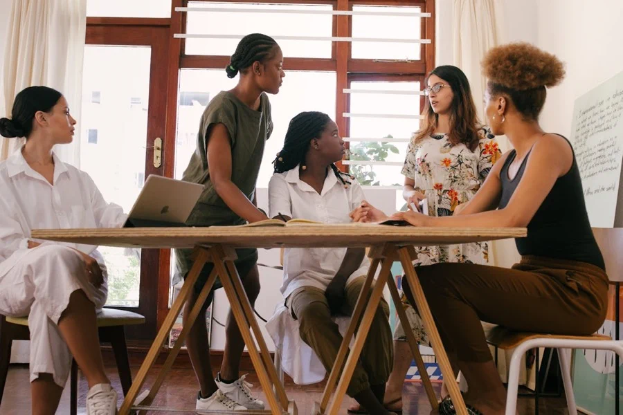 A diverse group of women engaged in a workshop session, seated around a table with notebooks and laptops
